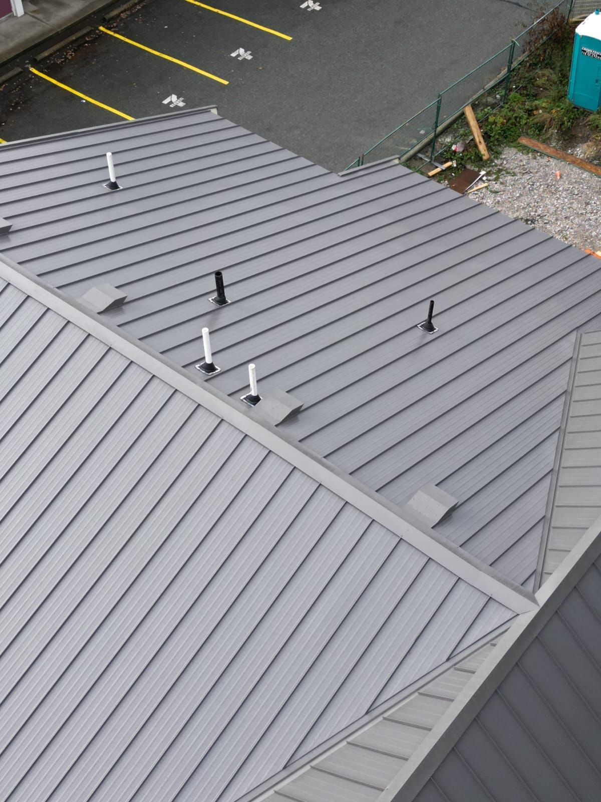 A high-angle view of a grey metal standing-seam roof with several plumbing vents, next to a parking lot.