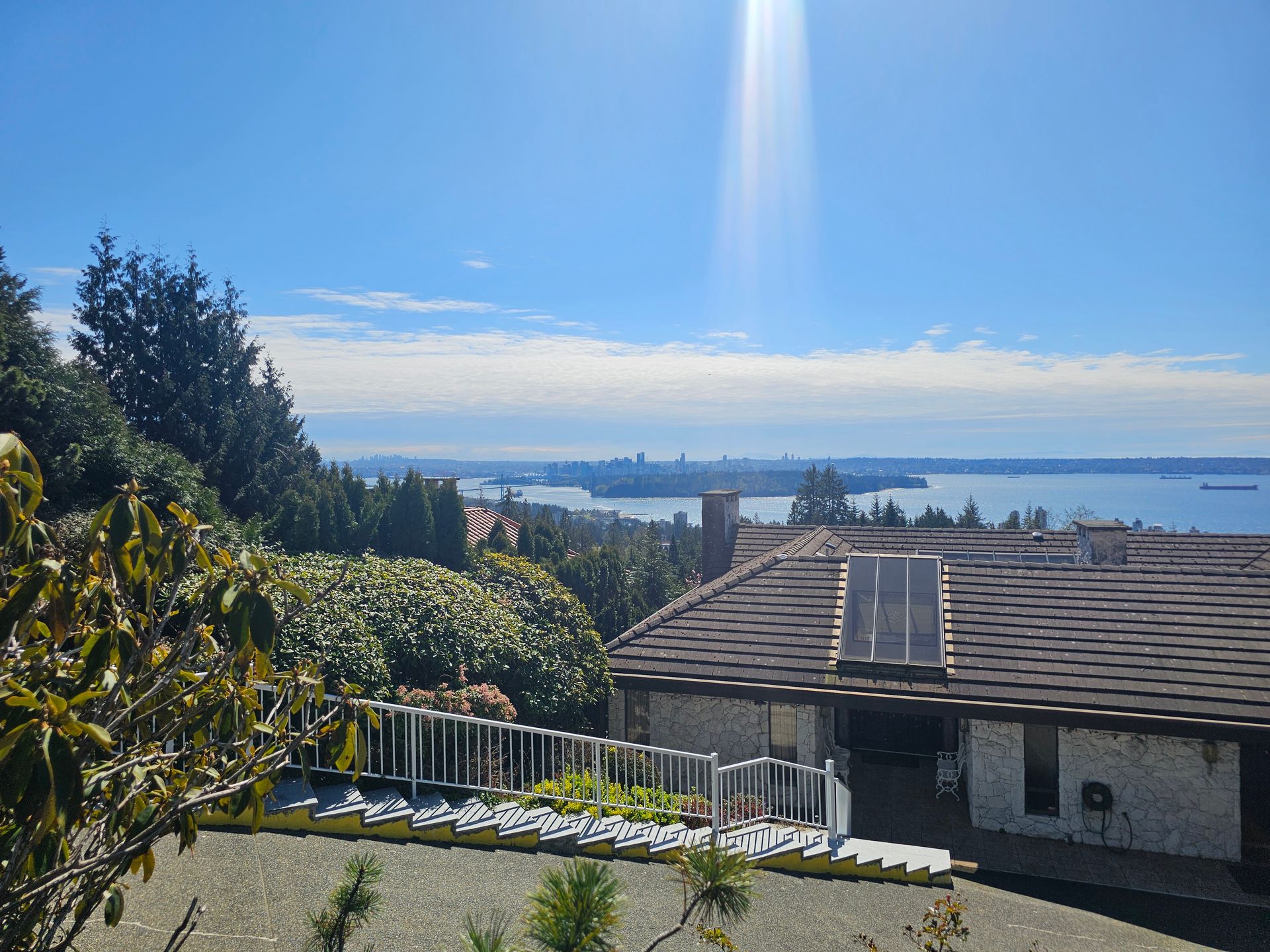 A scenic view of a house rooftop in the foreground, overlooking a bay with a distant city skyline under a sunny blue sky.