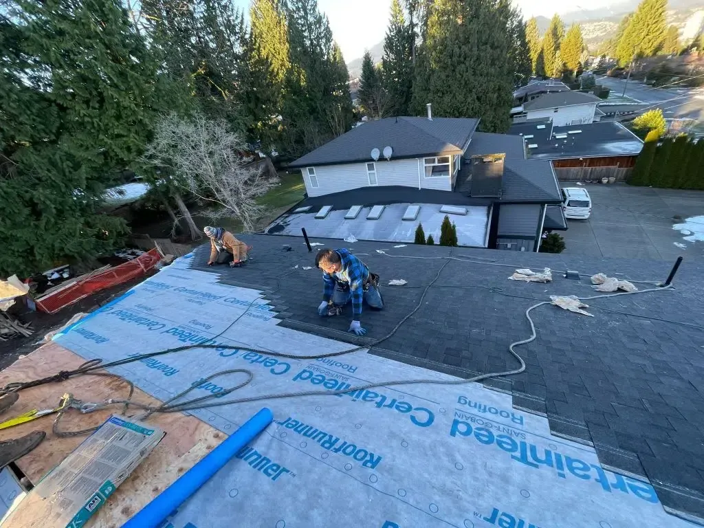 Two roofers install asphalt shingles over blue underlayment on a residential roof.