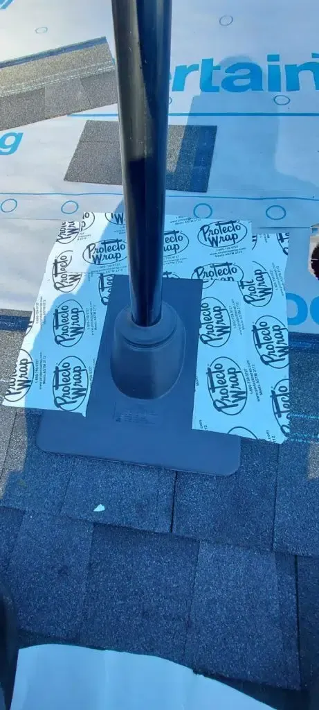 A black plumbing vent pipe installed on a roof, sealed with a gray flashing collar and branded adhesive flashing tape.