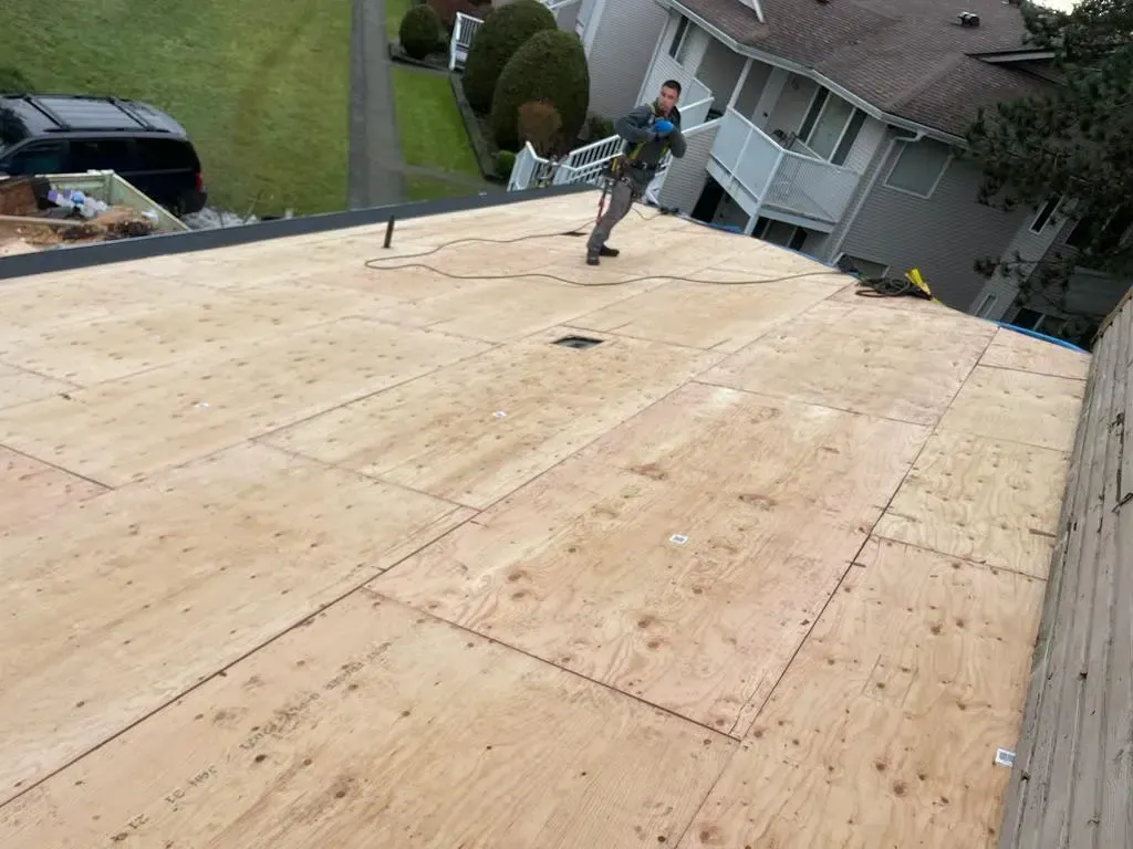 A construction worker stands on a plywood-sheathed roof during a residential roofing project.