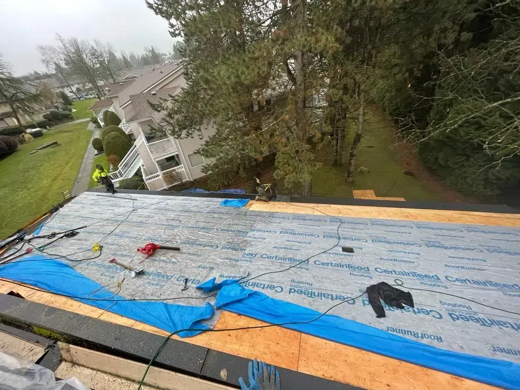 Workers on a residential roof installing light blue weather-resistant underlayment over plywood decking.