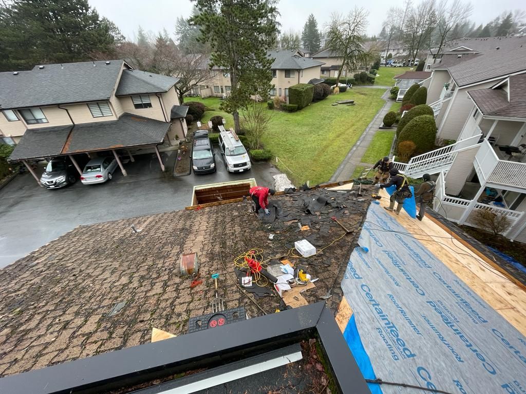 Workers repair a residential roof, installing new underlayment while removing old shingles, seen from an elevated angle.