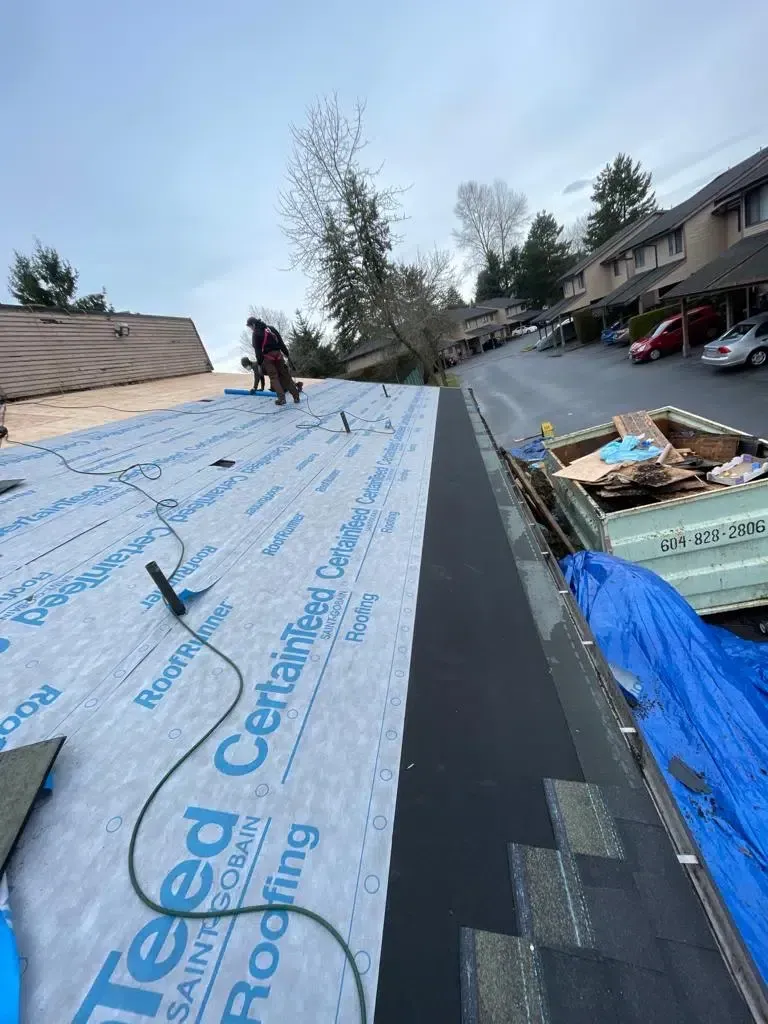 Roofing underlayment on a sloped roof with workers, shingles, and materials beside the house