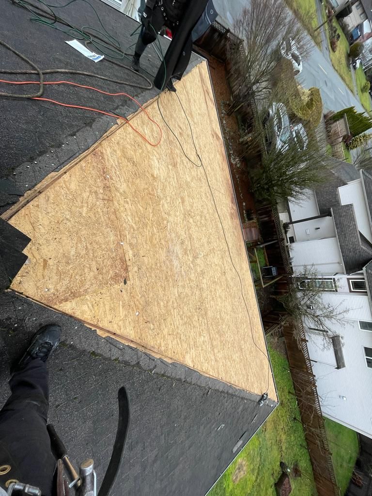 A worker stands on a partially reshingled roof, showing a large triangular area of exposed plywood sheathing.