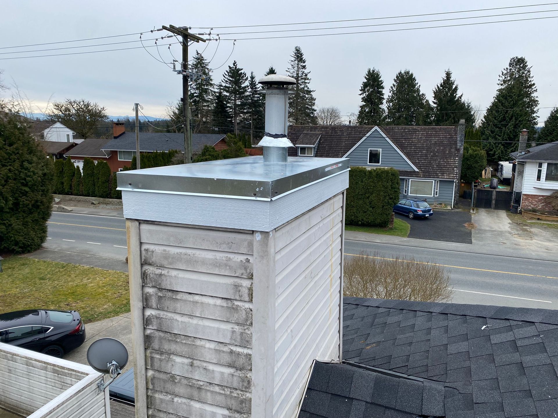A close-up view of a metal chimney cap atop a white-sided chimney on a shingled roof, with suburban houses in the background.