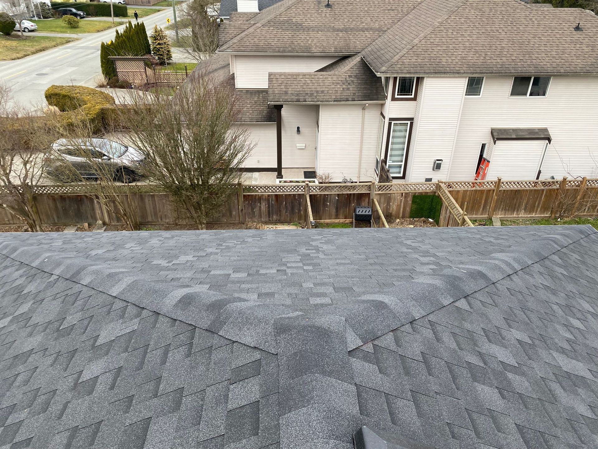 A high-angle view looking down at a gray asphalt shingle roof, with a white house and street visible in the background.