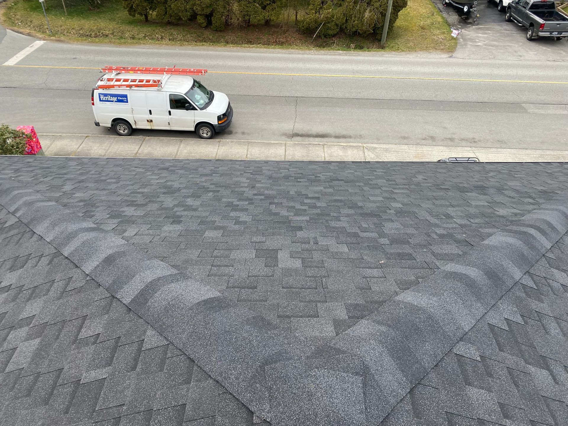 A high-angle view of a gray asphalt shingle roof, with a service van parked on the street below.