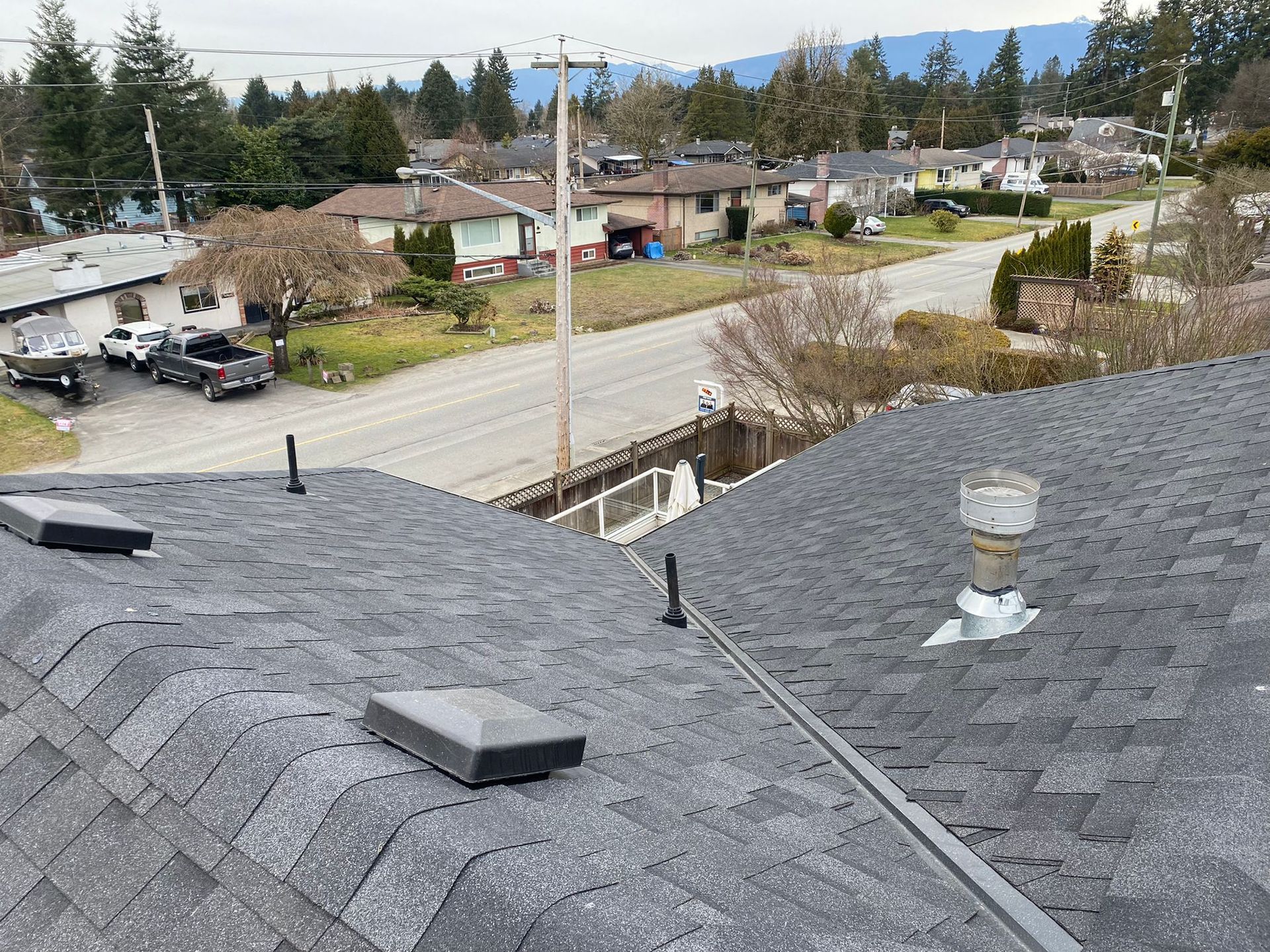 Rooftop shingles overlooking a suburban street with houses, trees, and distant mountains under a cloudy sky
