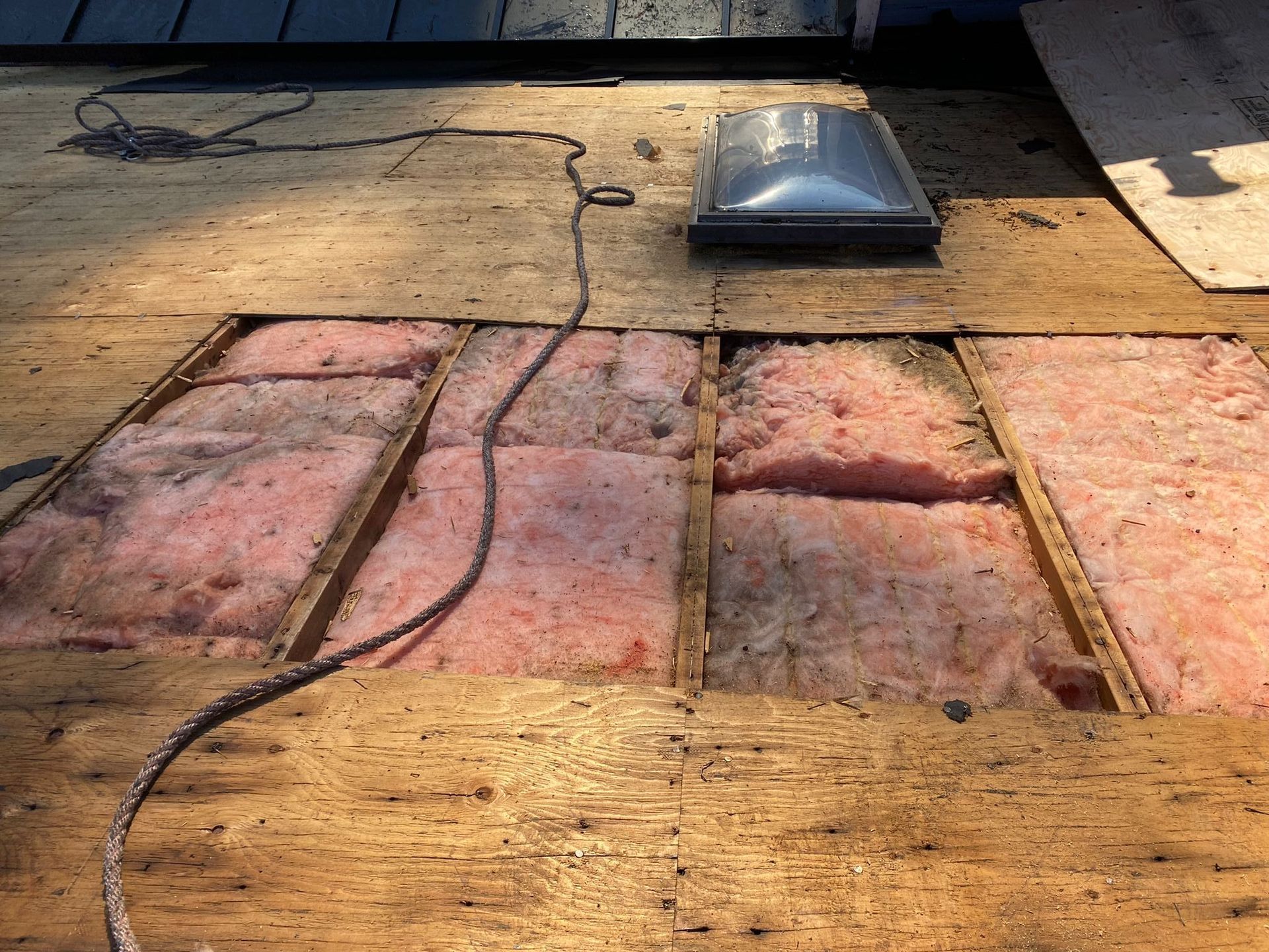 A section of a roof with plywood sheathing removed, revealing pink insulation between wooden joists and a skylight nearby.