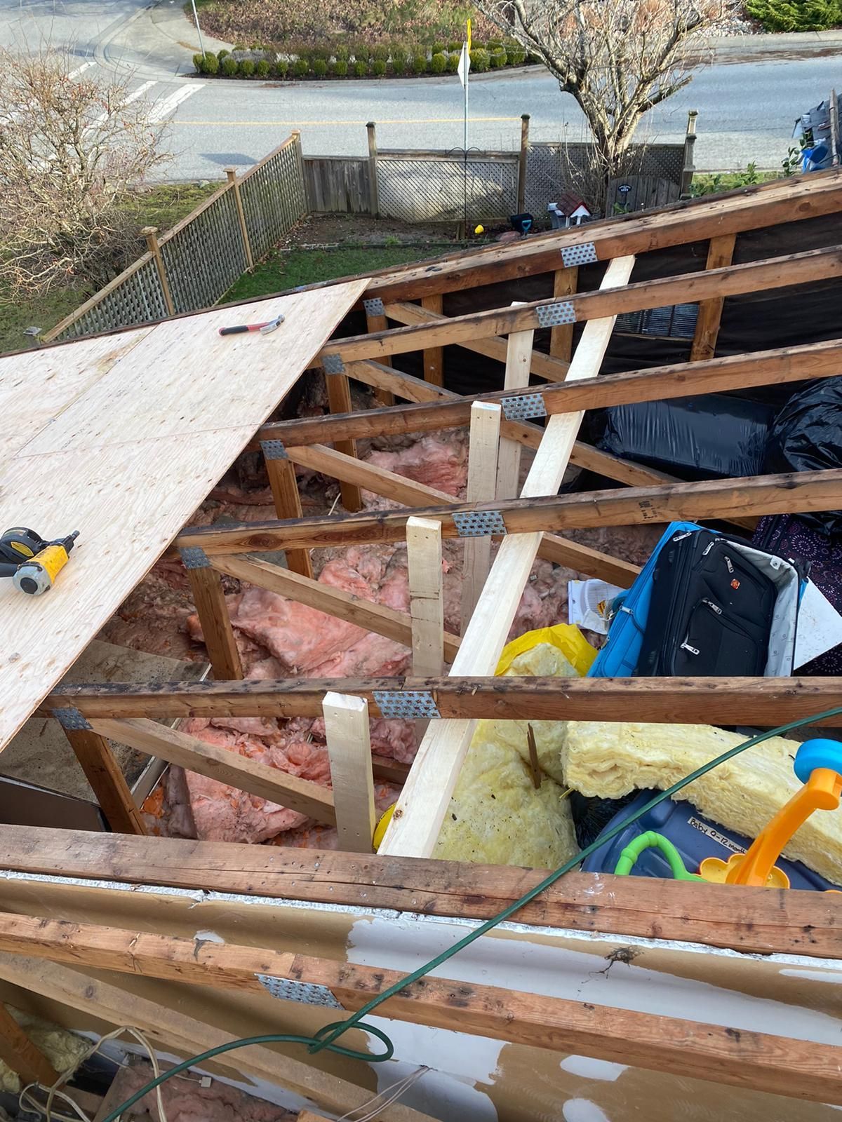 A partially exposed residential roof under repair, showing wooden trusses, insulation, and a sheet of plywood being installed.
