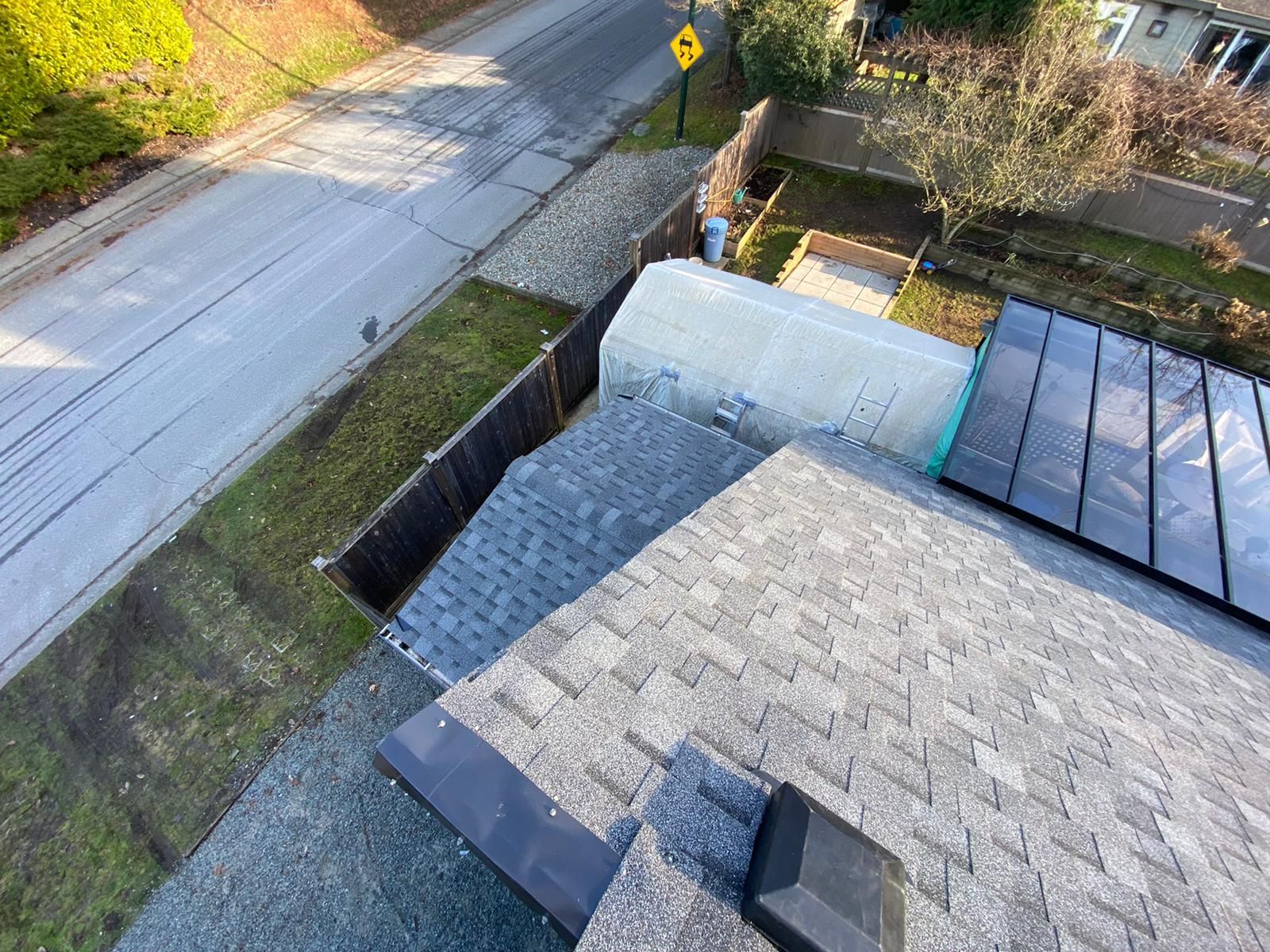 An elevated view of a gray shingled roof, a dark solar panel array, and a white shed, next to a street and fence.