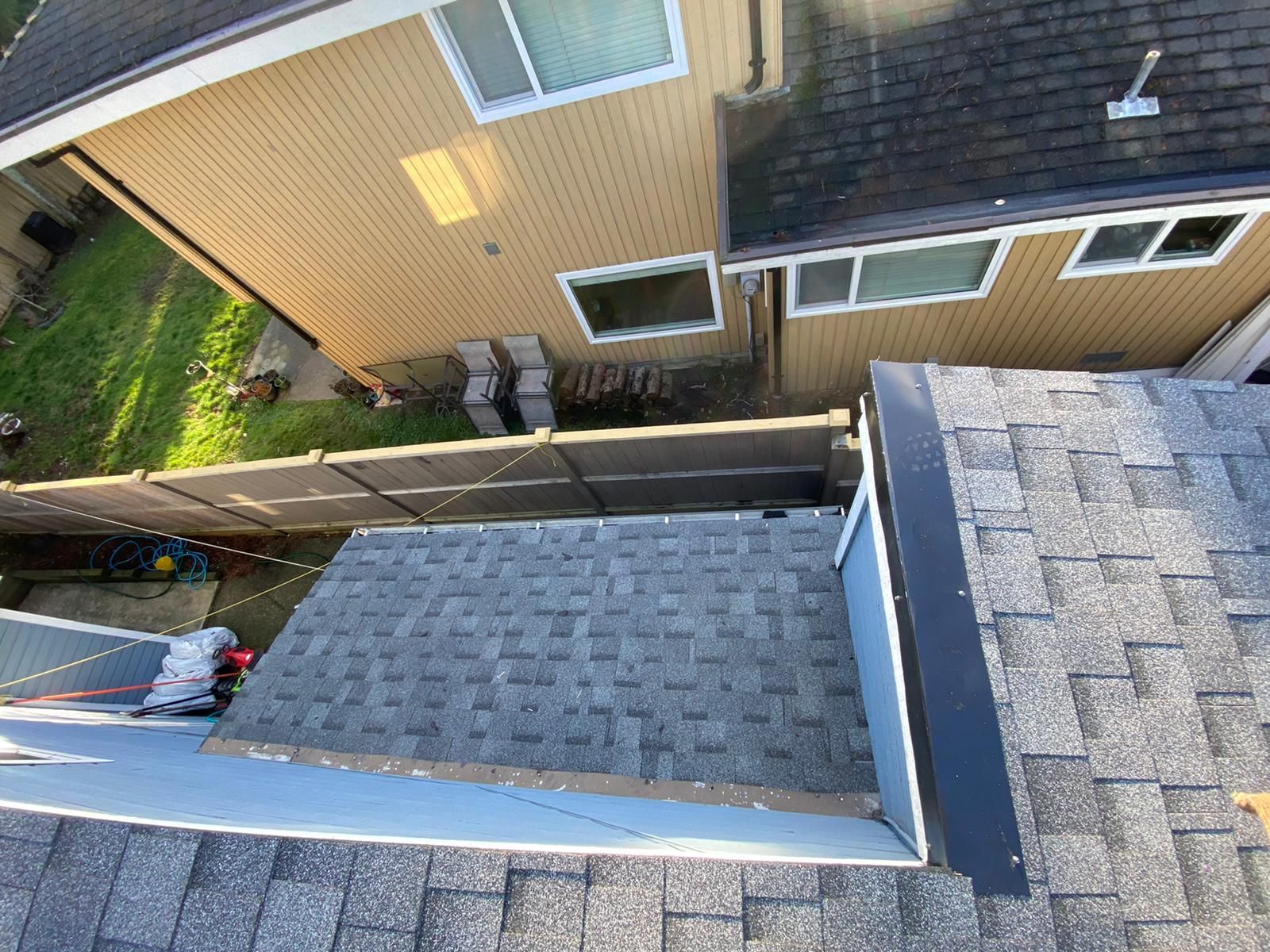 An elevated view of shingled roofs on a residential building, with a neighbor's house and fence visible in the background.