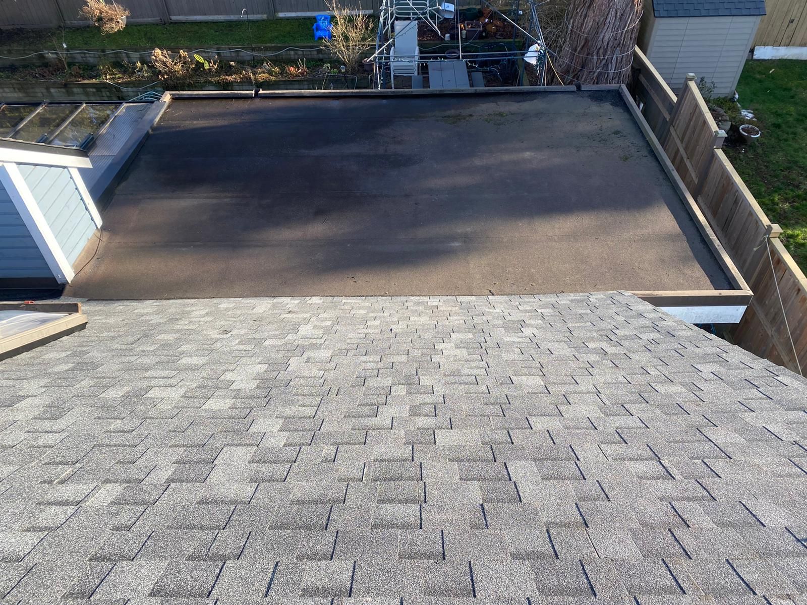 A view from a shingled roof looking down onto a rectangular, flat black roof surrounded by a wooden fence.