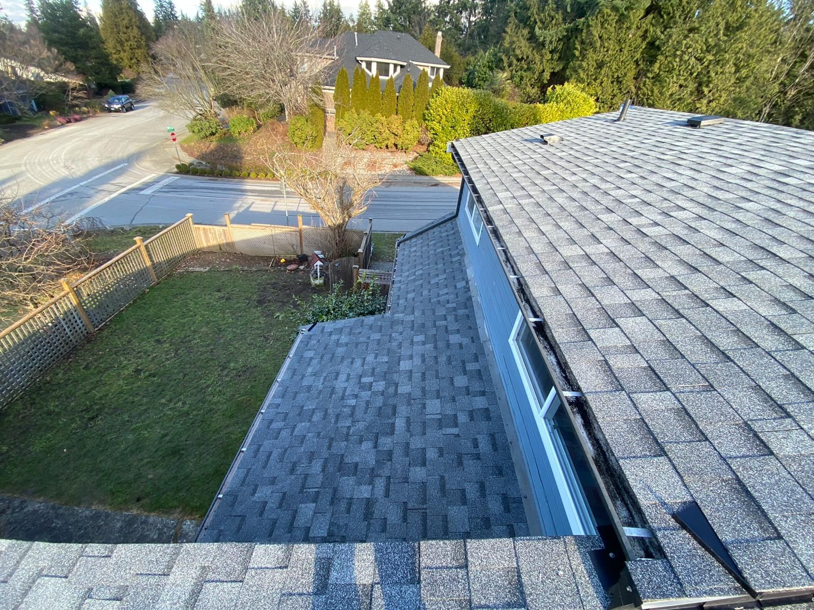 A high-angle view of a gray shingled roof extending toward a residential street, a grassy yard, and surrounding trees.