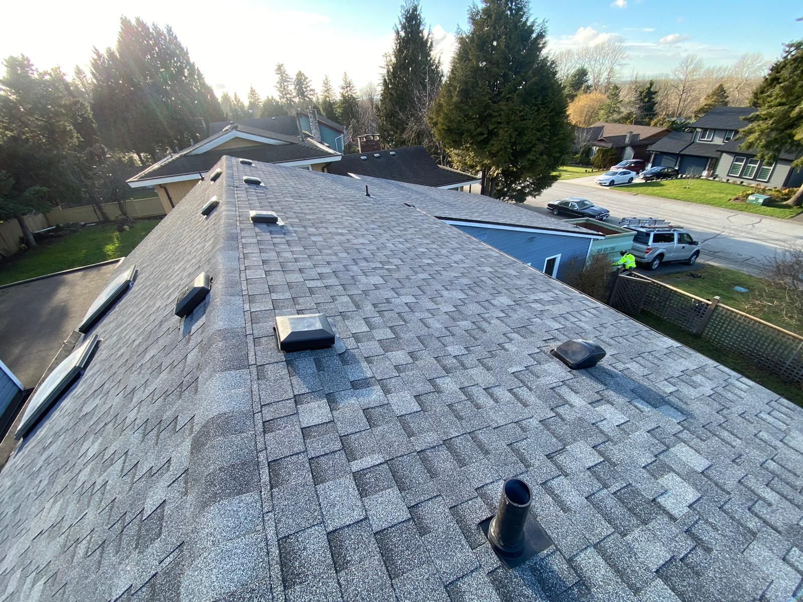 A high-angle view of a gray shingled roof with several black vents and a plumbing stack, overlooking a suburban street.