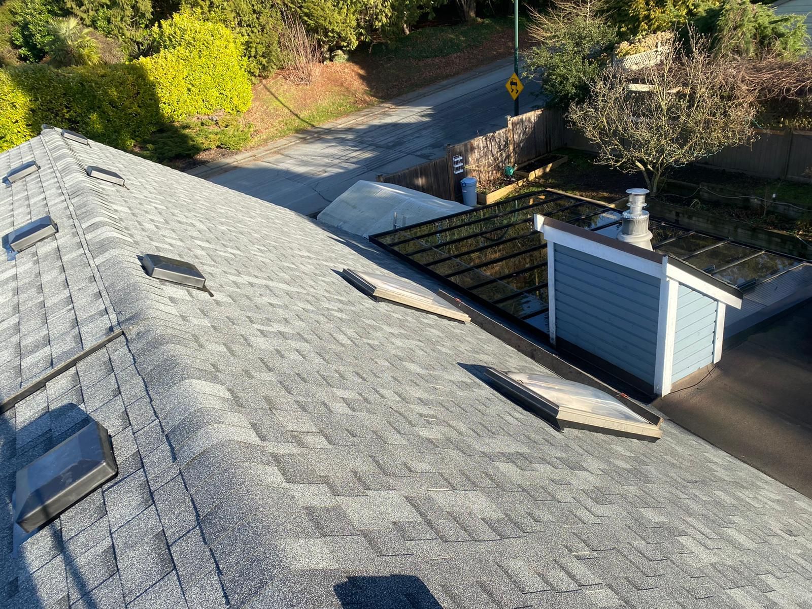 An overhead view of a gray shingled roof with four skylights, next to a blue structure and a tree-lined street.