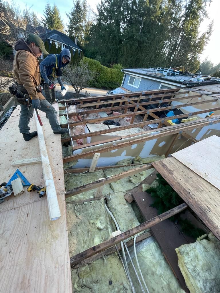 Two workers stand on the plywood roof of a residential home under renovation, exposing the interior framing and insulation.