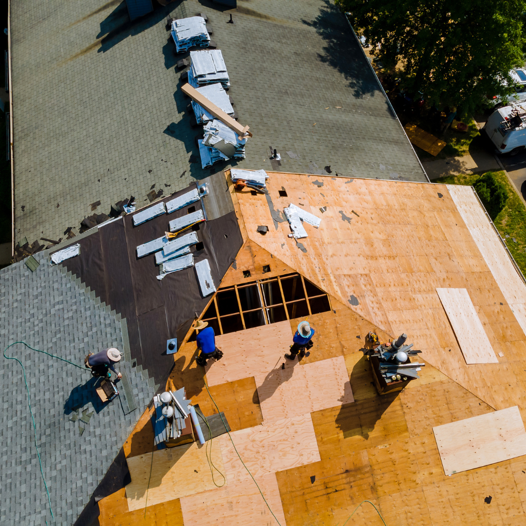 Aerial view of workers replacing shingles on a house roof, showing exposed wood decking and stacks of construction materials.