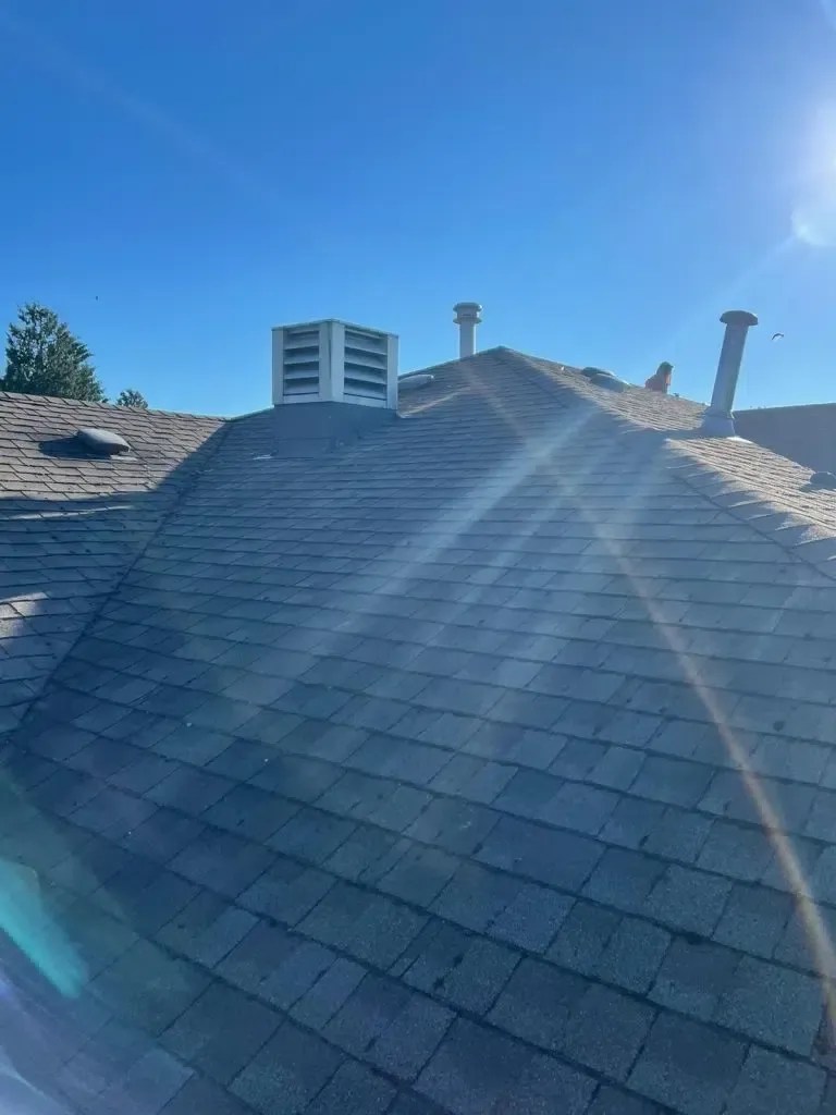 A close-up view of a shingled roof under a clear blue sky, featuring a white box vent and two metal chimney pipes.