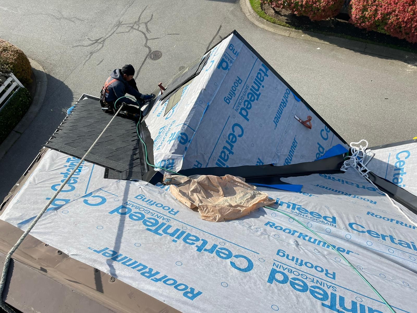 A worker in protective gear repairs a residential roof covered in blue and white underlayment, secured by a safety rope.