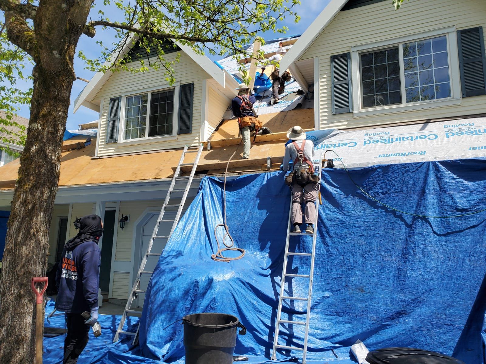 Workers on ladders repairing a house roof, with blue tarps covering the front wall and debris below.