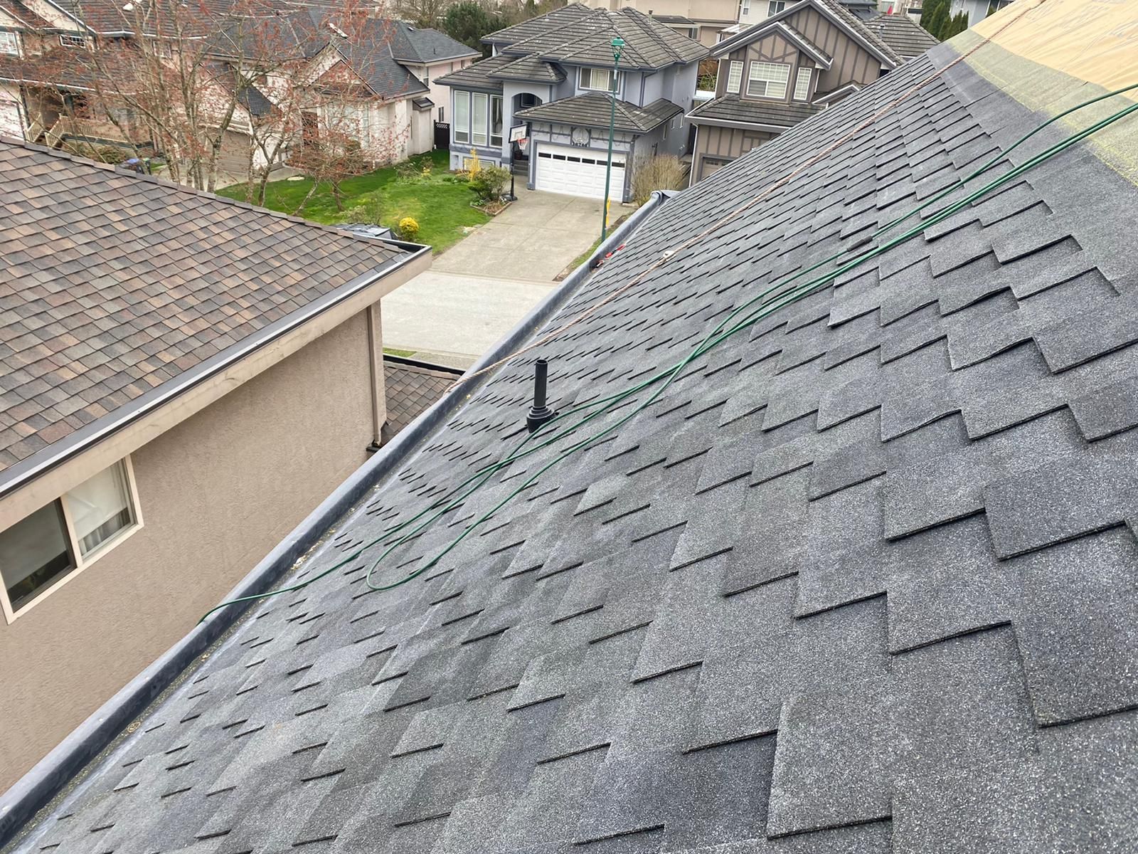 A view from a roof showing gray asphalt shingles, a black vent pipe, and a neighboring house in a residential area.