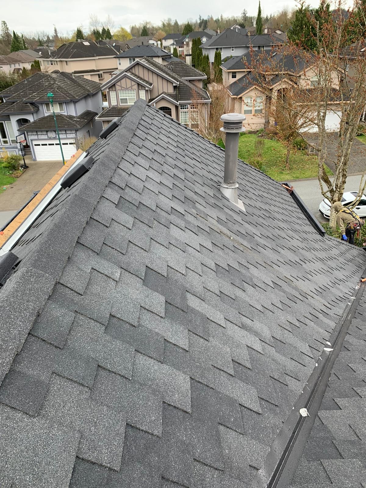 A high-angle view of a grey shingled roof with a chimney, overlooking a residential neighborhood with houses and trees.
