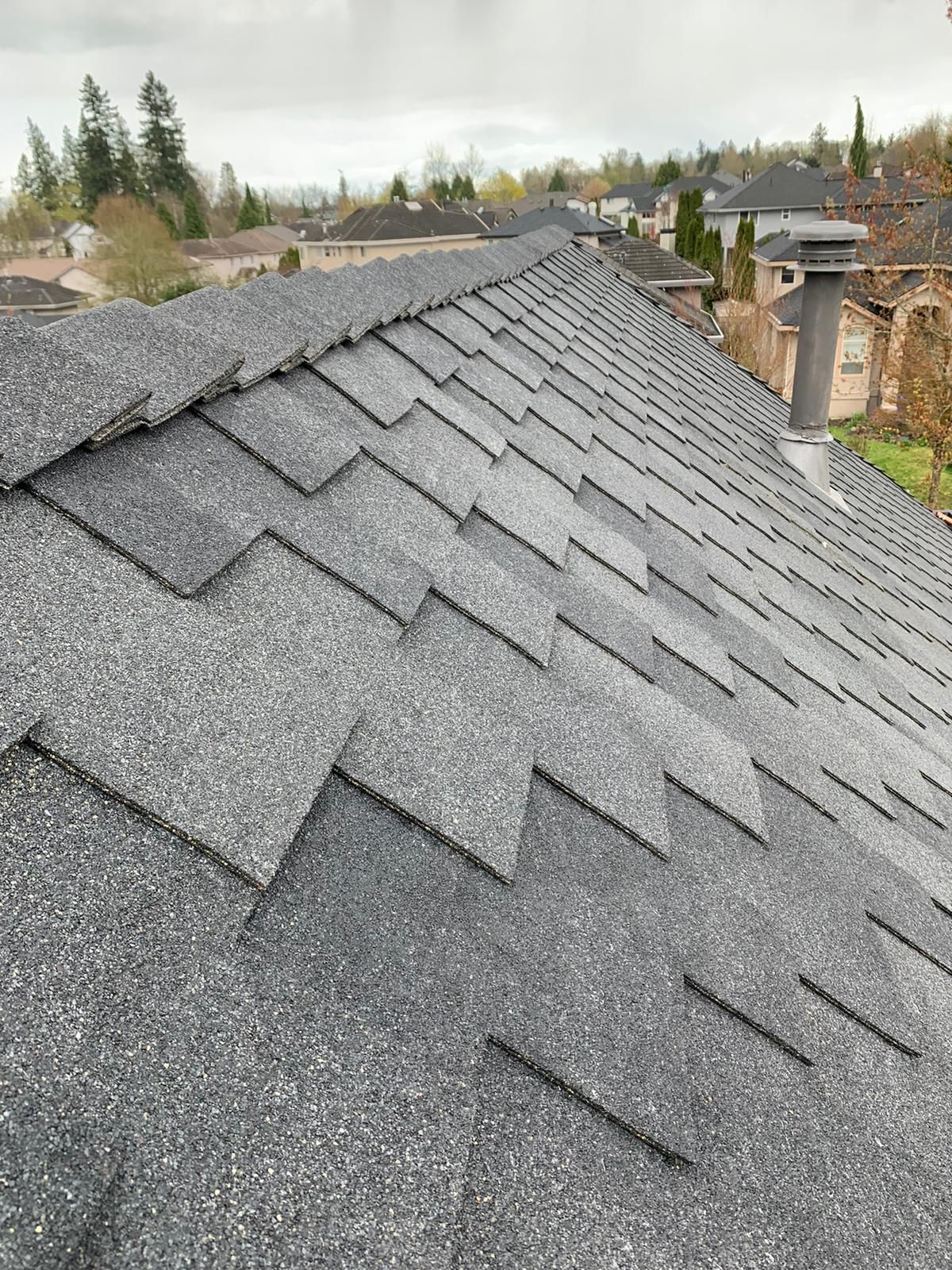 A view looking down the slope of a gray shingle roof toward a suburban neighborhood on an overcast day.
