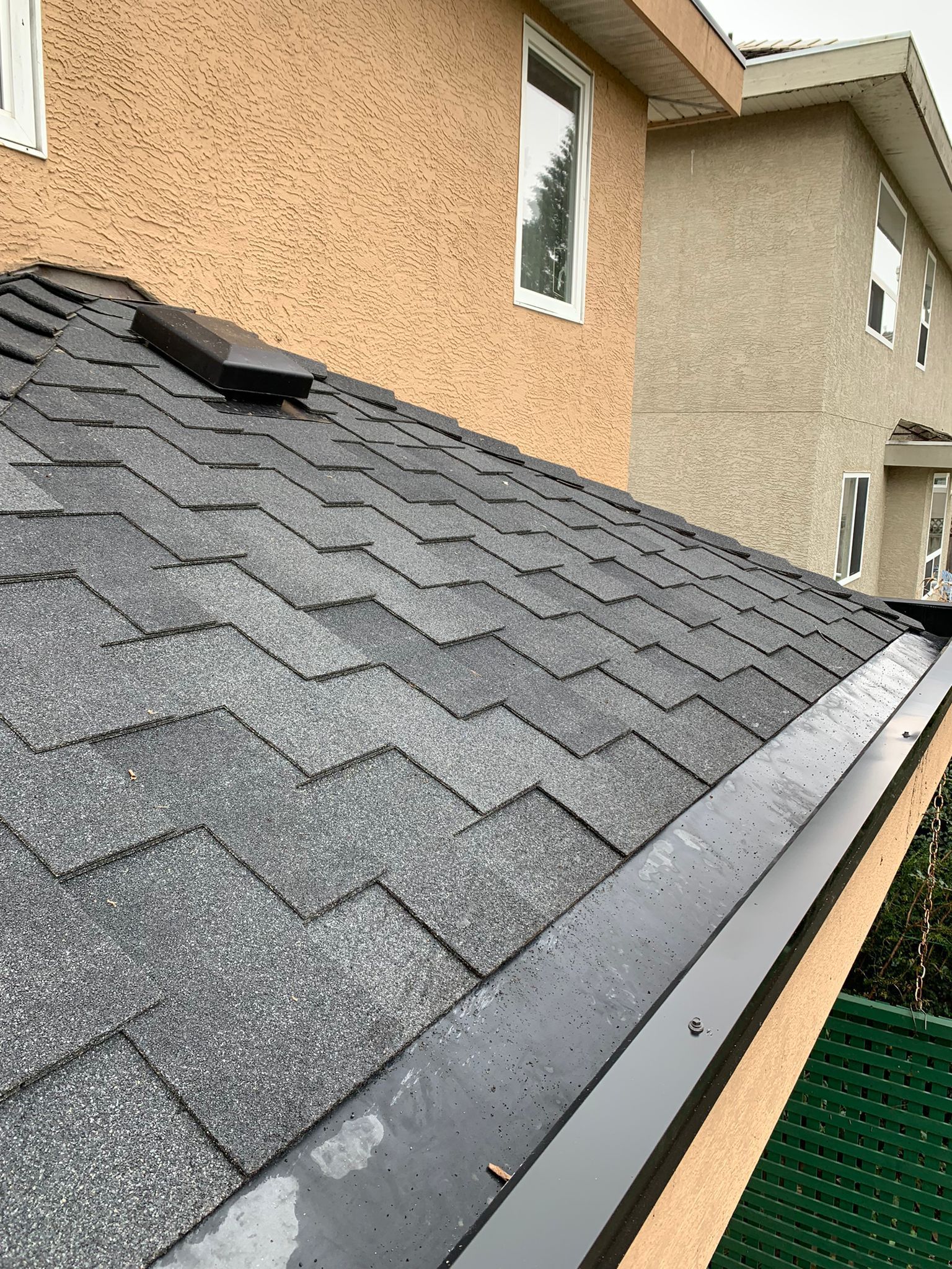 A close-up of a gray shingled roof with a black vent on a beige stucco house with a gutter visible at the edge.