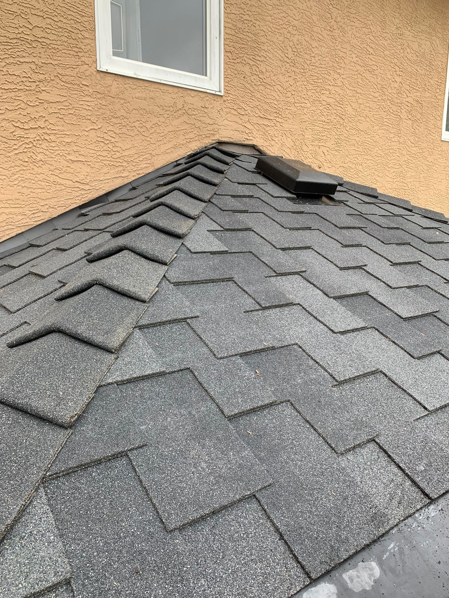 A close-up view of a shingled roof slope with a black ventilation cap, set against a tan, textured exterior wall.