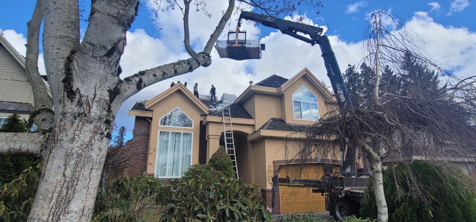 A crane with a hanging basket lifts workers toward the roof of a beige house to perform repairs.