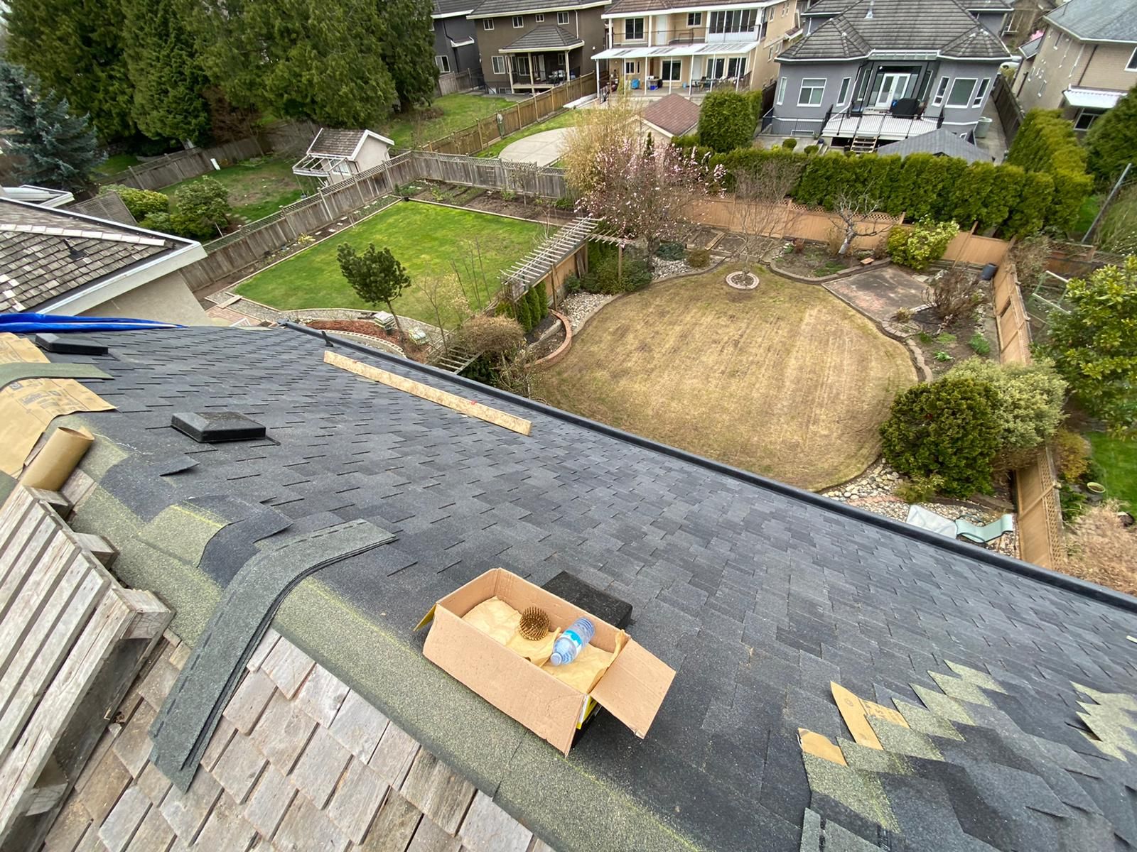 A view from a partially shingled residential roof overlooking a backyard with a circular lawn and neighboring houses.