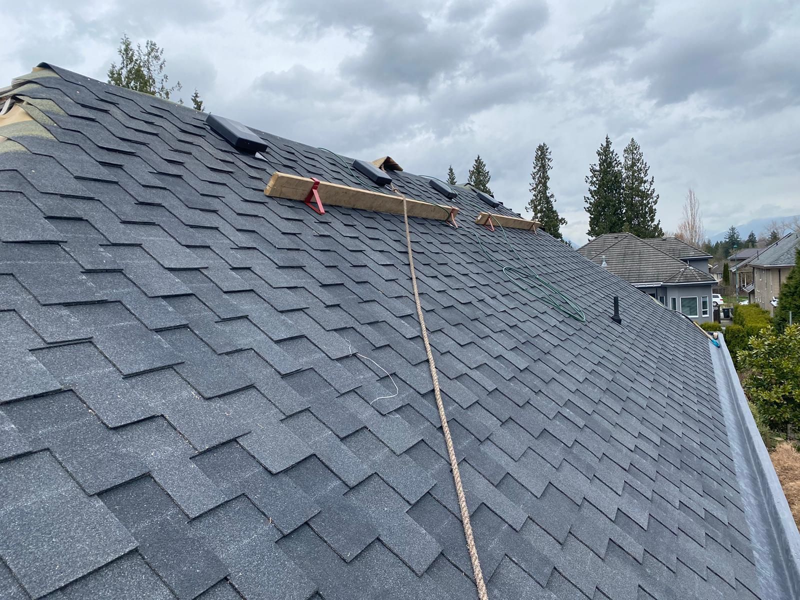 A view of a shingled roof with a safety rope, wood board, and metal vents, under a cloudy sky.