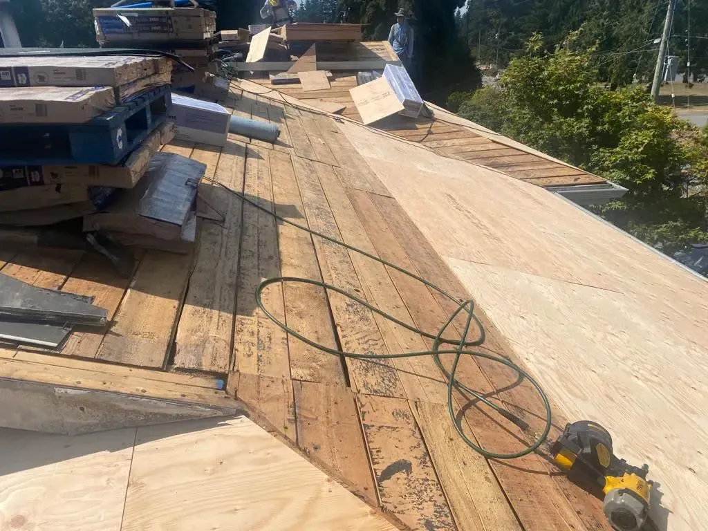 A residential roof under construction with wooden decking, a stack of materials, and a nail gun with its cord.