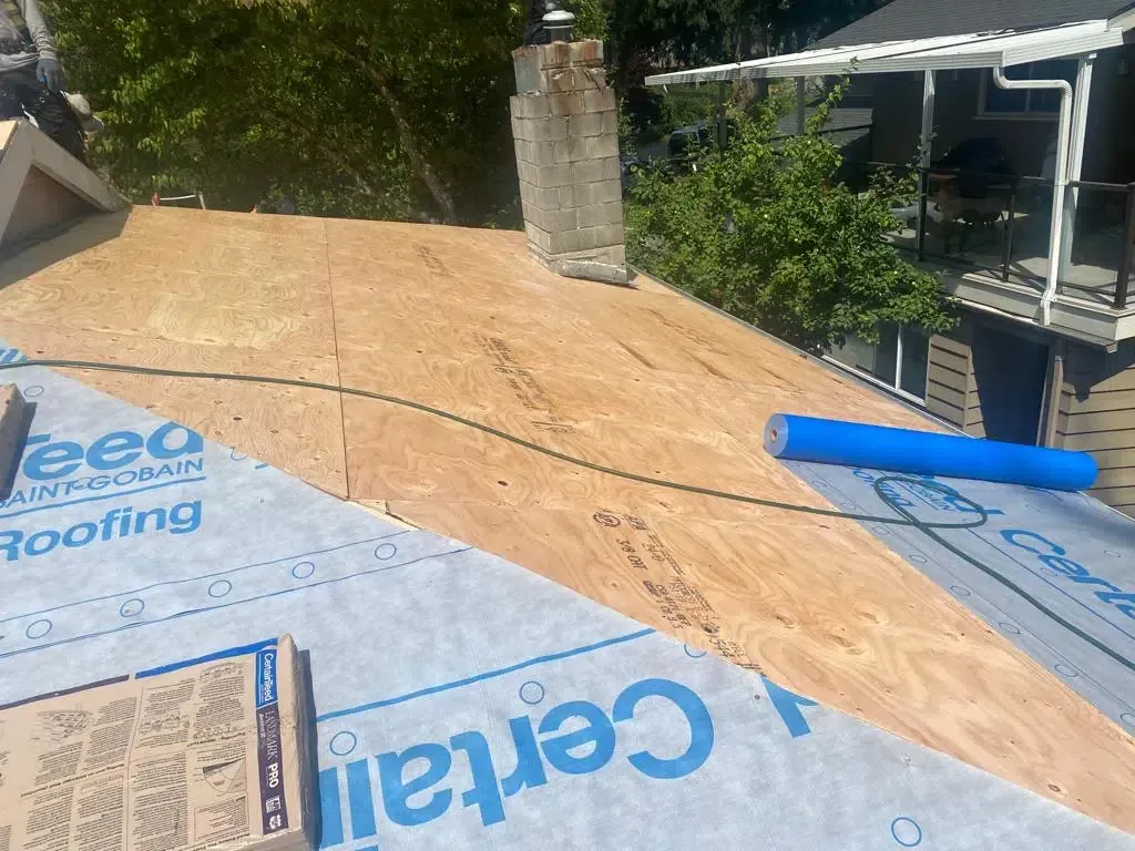 A roof under construction with plywood decking, a brick chimney, and blue CertainTeed synthetic underlayment.