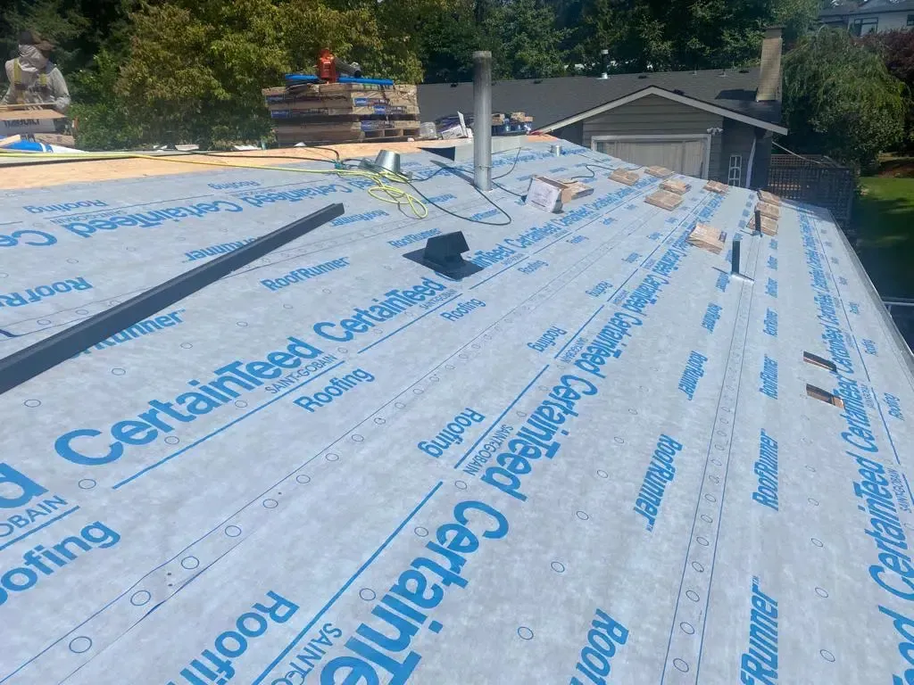 A roof under construction covered with light blue CertainTeed underlayment, with a worker visible in the background.