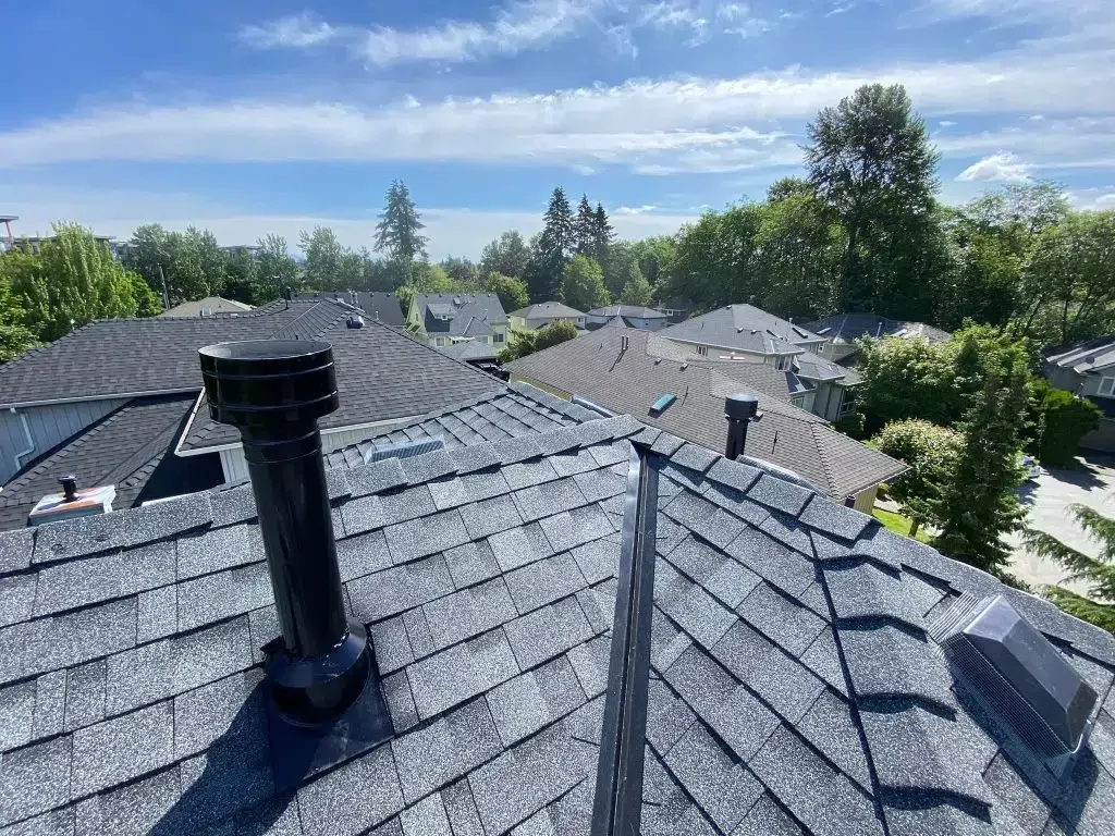 A high-angle view of a gray asphalt shingle roof featuring a black chimney cap and a roof vent under a bright blue sky.