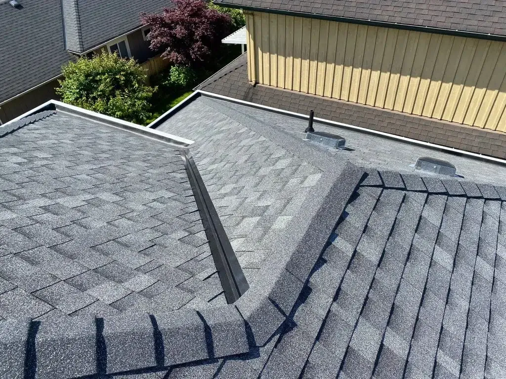 An elevated, angled view of gray shingled roof sections, with a black flashing strip and a yellow-walled building behind.