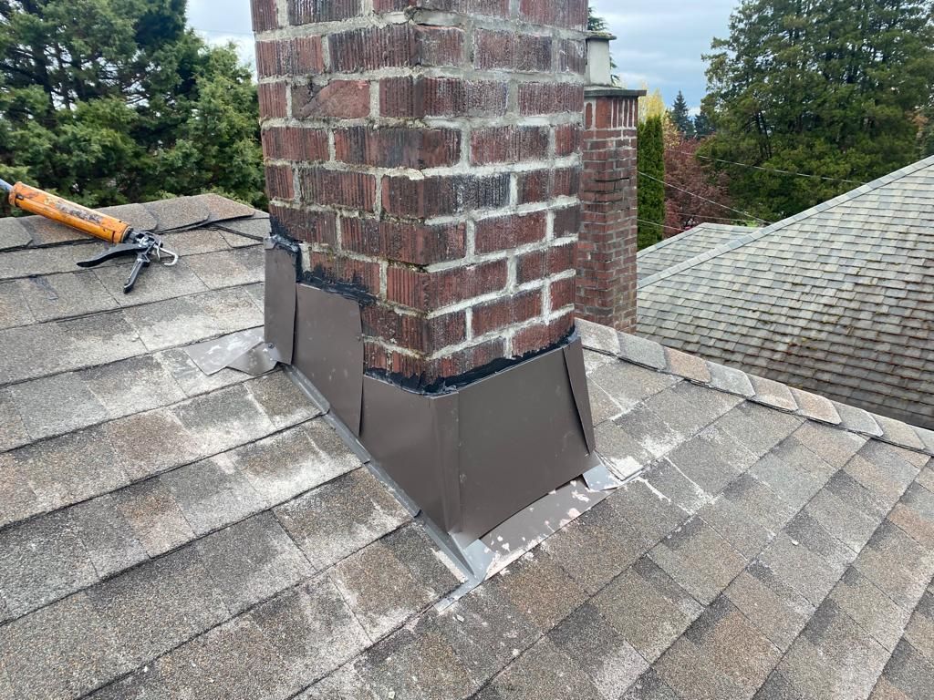 New brown metal flashing installed around the base of a brick chimney on a shingled roof.