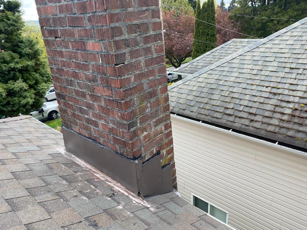 A brick chimney on a shingled roof with dark metal flashing at the base, viewed from above near a neighboring house.
