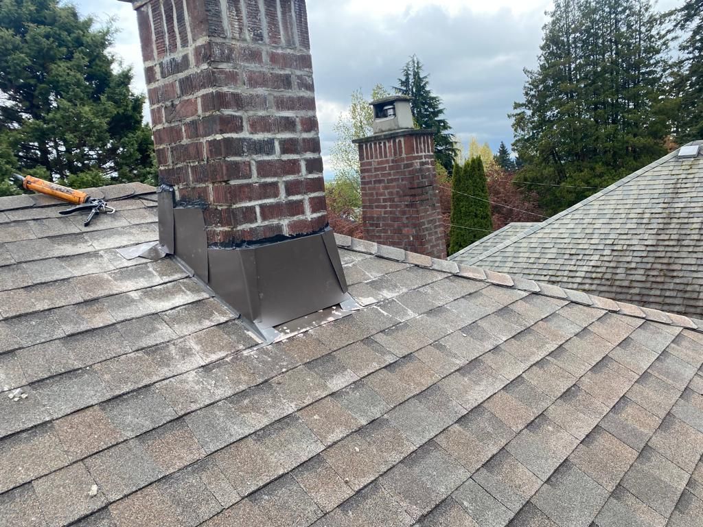 A brick chimney with metal flashing on a shingled roof, with another chimney and trees visible in the background.