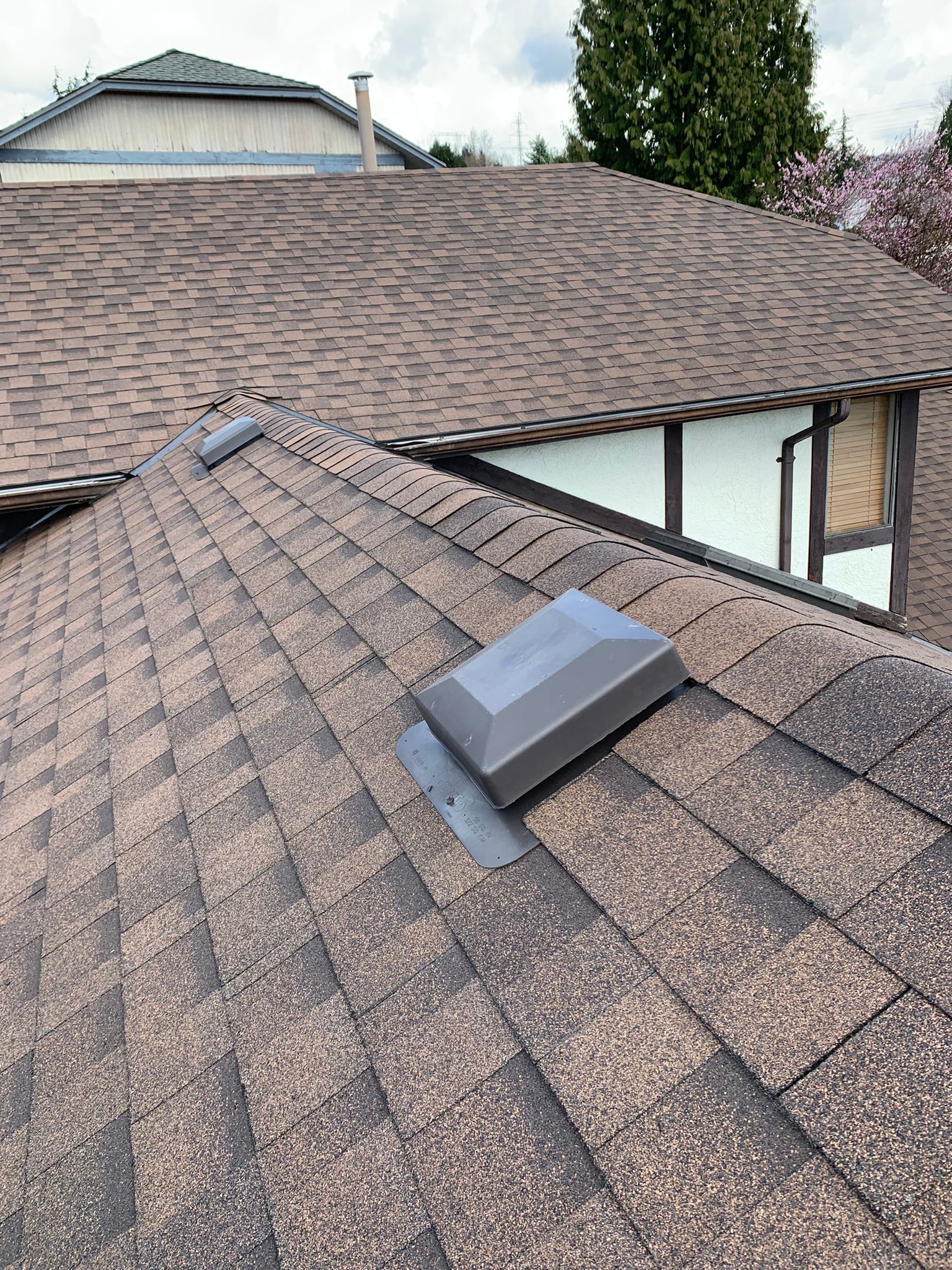 A view of a shingled roof with two dark grey ventilation hoods installed on the slope, with another roof visible behind.
