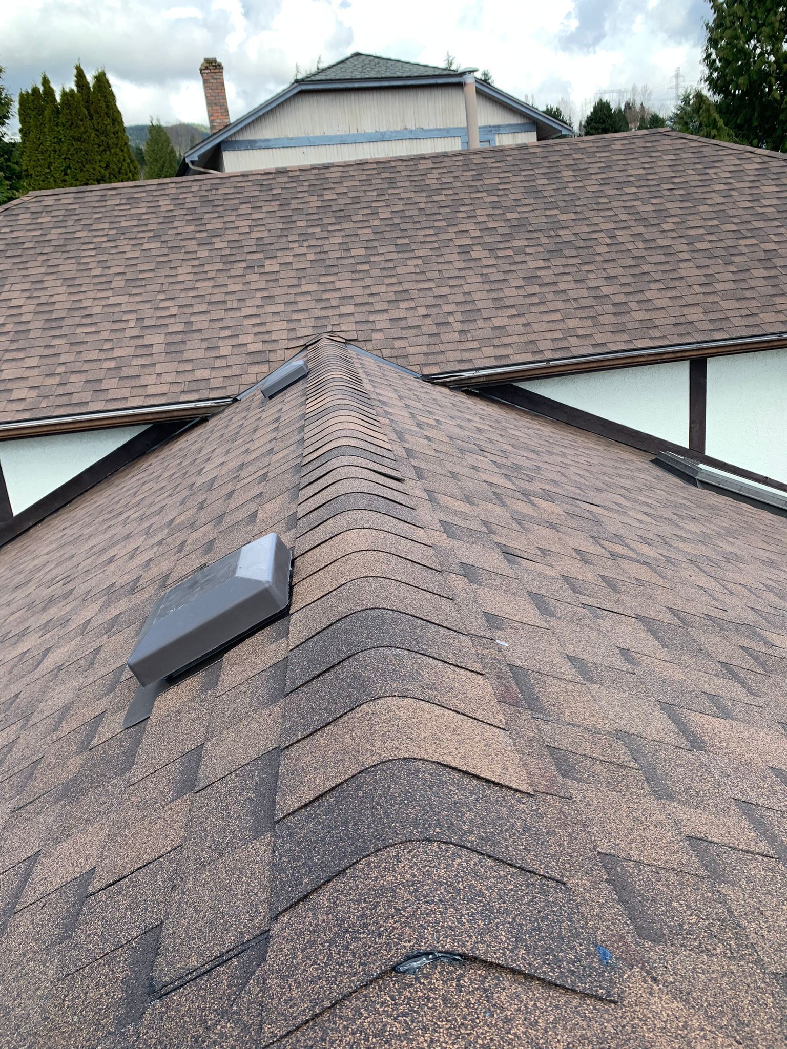 A high-angle view of a brown shingled roof with a ridge vent and a grey square ventilation fan.
