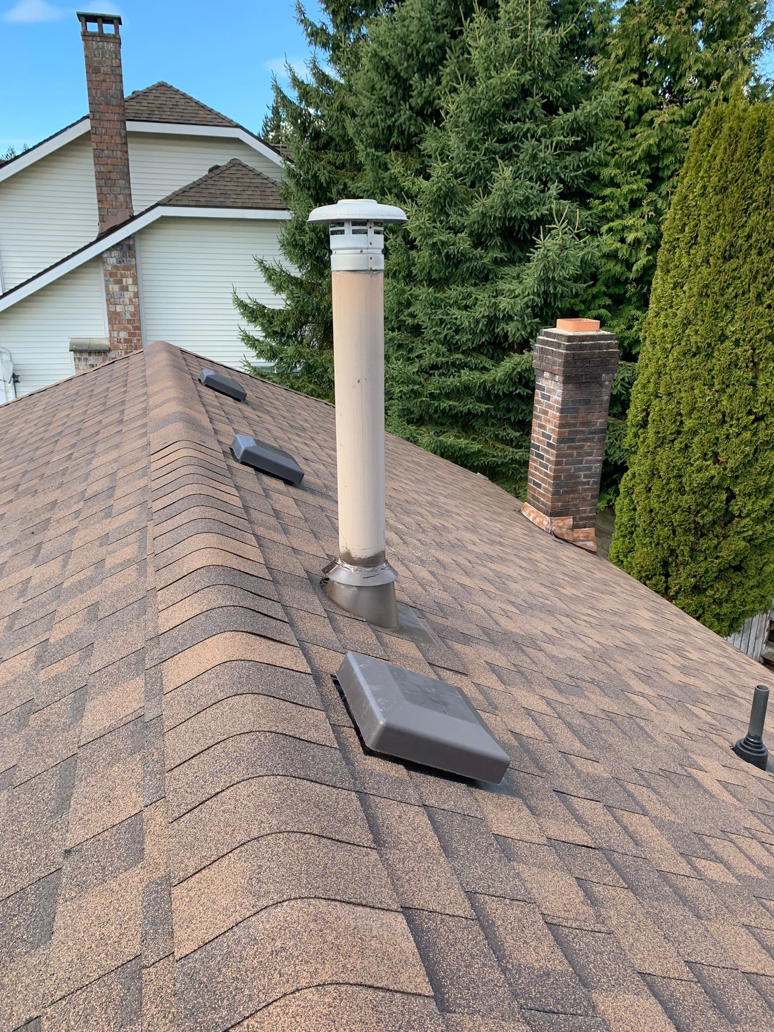 A brown shingled roof features a central beige exhaust vent pipe, several square roof vents, and a brick chimney.