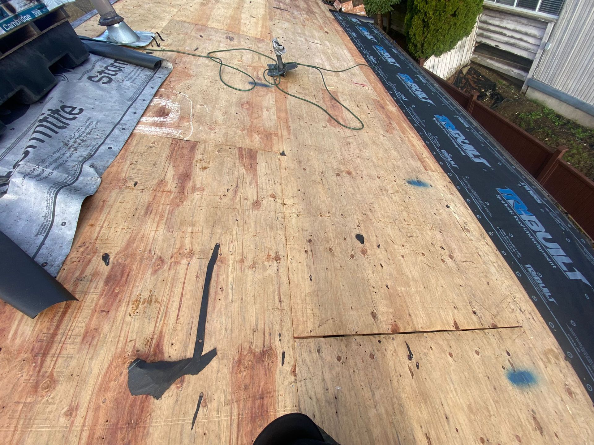 An unfinished residential roof featuring bare plywood decking and rolls of black synthetic roofing underlayment.