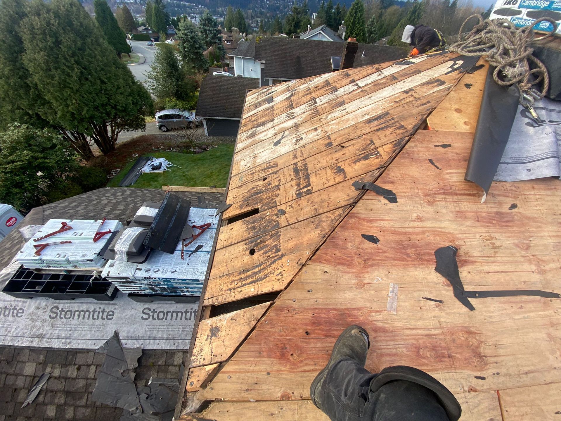 A roof under construction showing exposed plywood sheathing, roofing materials, and a worker's leg in the foreground.