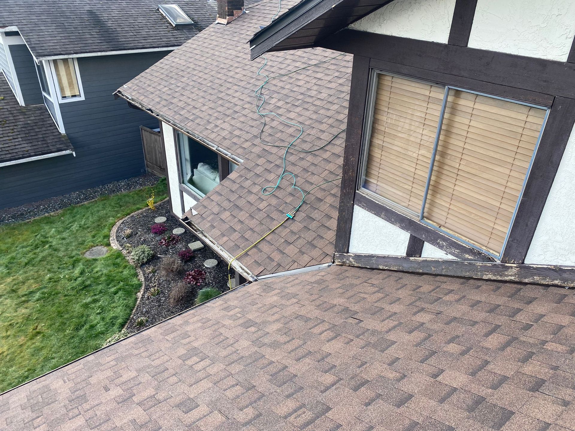 High-angle view of a brown shingled roof with a string of holiday lights, adjacent to a house with a Tudor-style window.