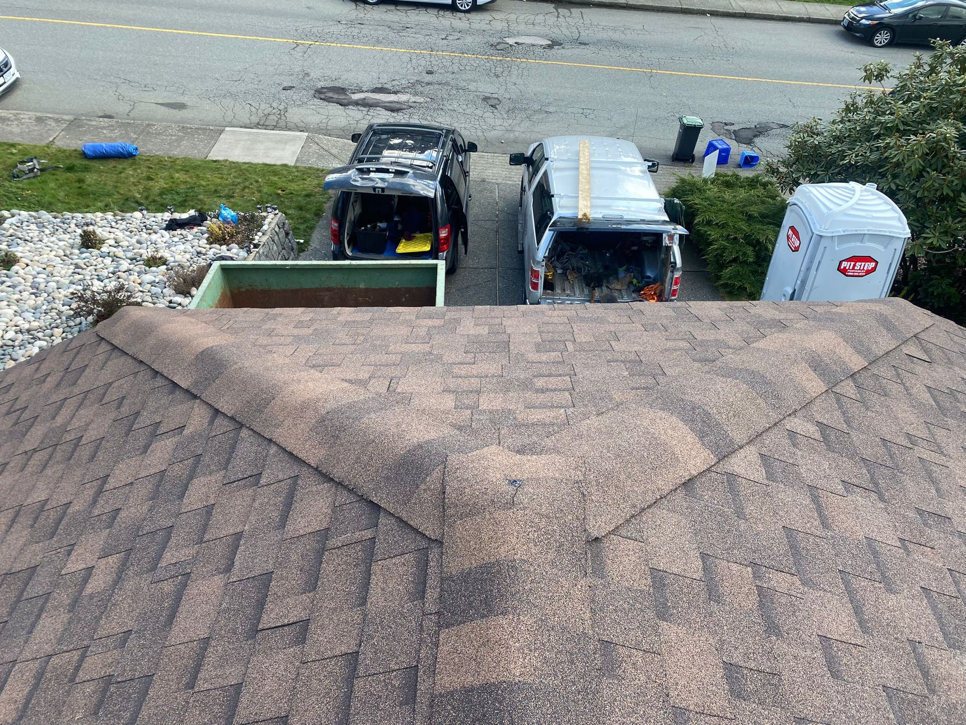 A view from a shingled roof looking down at a driveway with two cars, a dumpster, and a portable toilet.