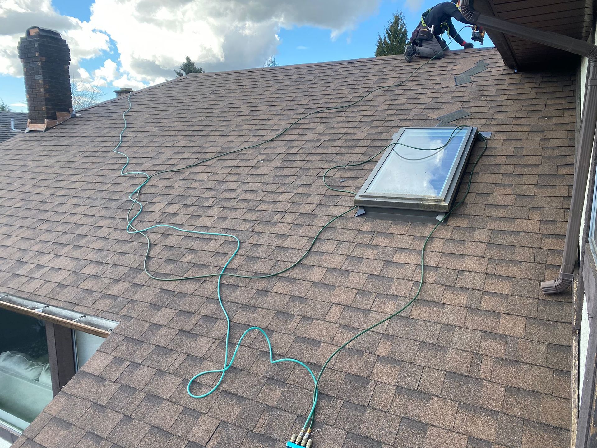 A worker kneels on a brown shingled roof next to a skylight with a green hose coiled nearby.